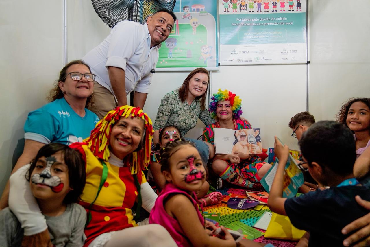 A foto mostra a primeira-dama, profissionais da gestão e crianças reunidos em uma sala durante uma atividade de incentivo à leitura. Uma mulher vestida de Emília segura um livro aberto, mostrando ele para as crianças